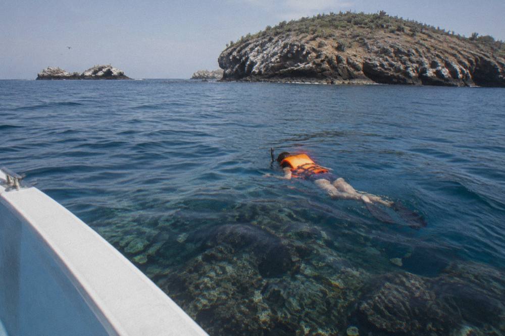 Snorkeler in orange life jacket swimming beside a boat near rocky islands