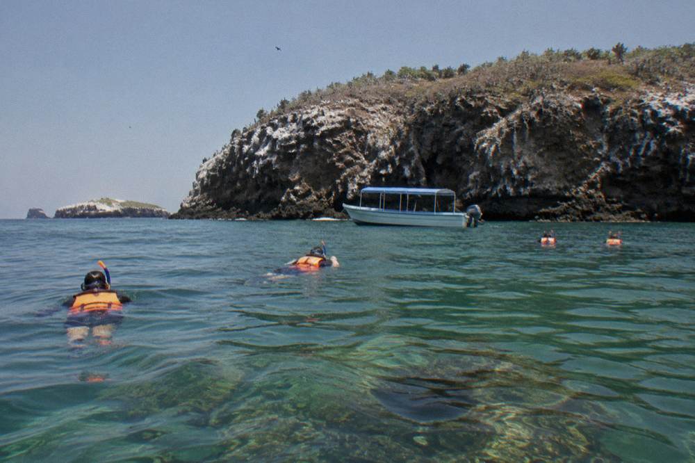 A covered small boat anchored near rocky islands with snorkelers in the water beside it