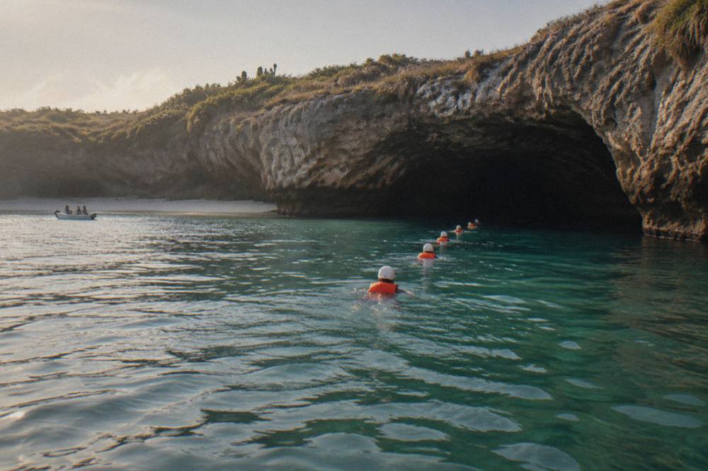 Swimmers in orange life jackets swimming toward a sandy beach beneath a rocky arch