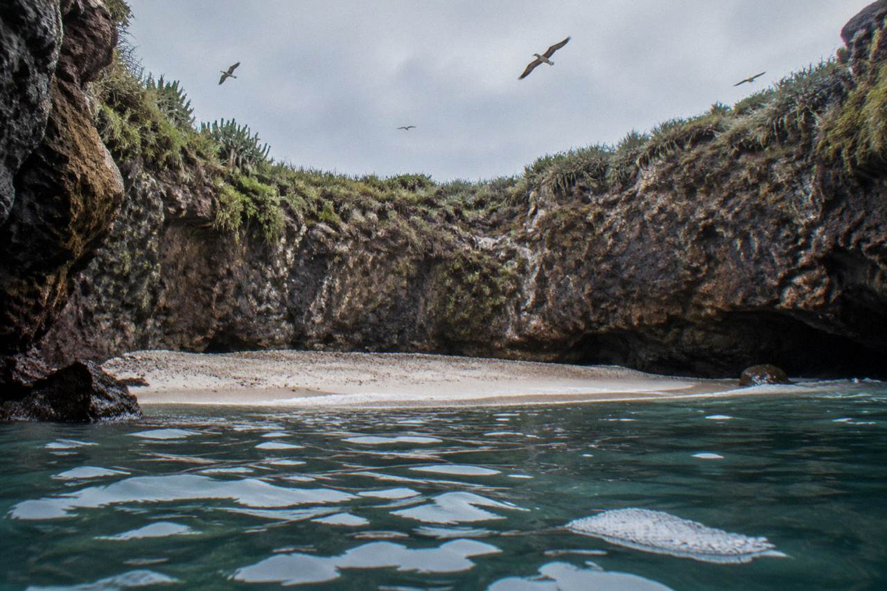 A crater beach seen from the water inside the Marietas Islands, with rock walls rising around a narrow sandy cove