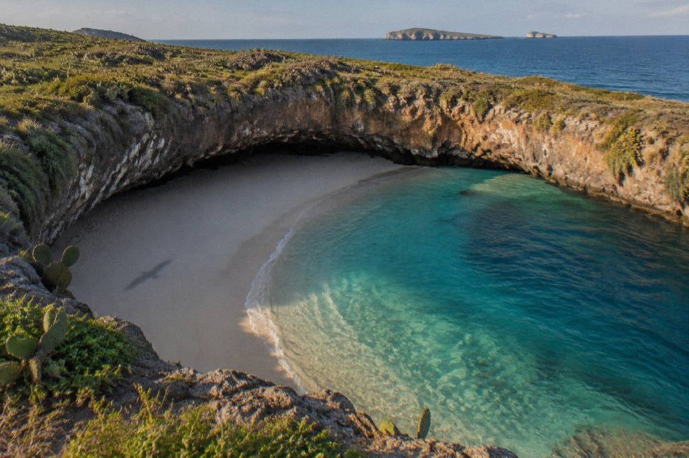Sandy beach inside a rocky crater with clear turquoise water and distant islands