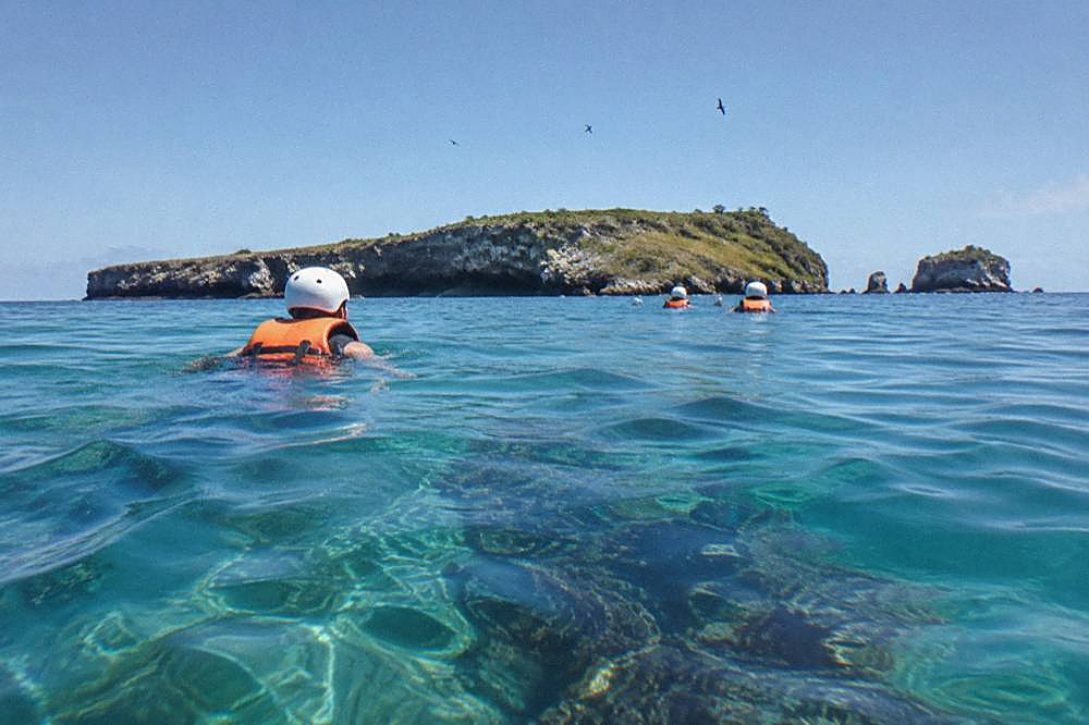 People in helmets and life jackets swimming in clear turquoise water toward a rocky island