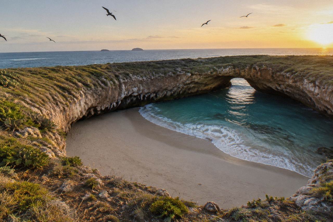 A sandy beach inside a rocky crater open to the sky, with turquoise water at the entrance passage