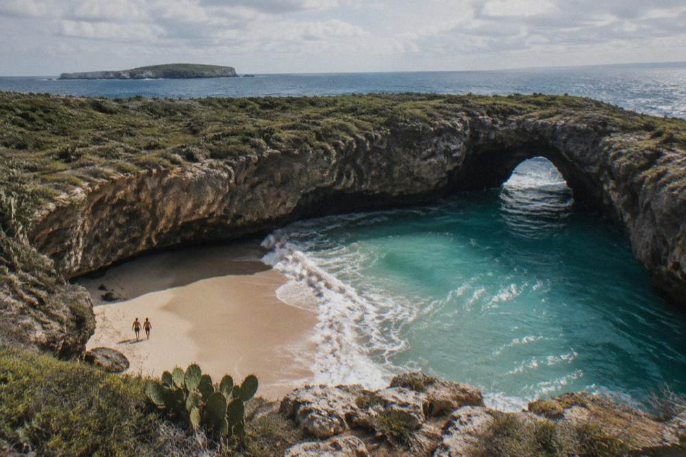 Two people standing on a crater beach enclosed by cliffs with a sea arch