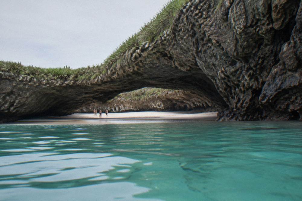 Rocky arch opening onto a sandy crater beach with clear turquoise water and distant figures