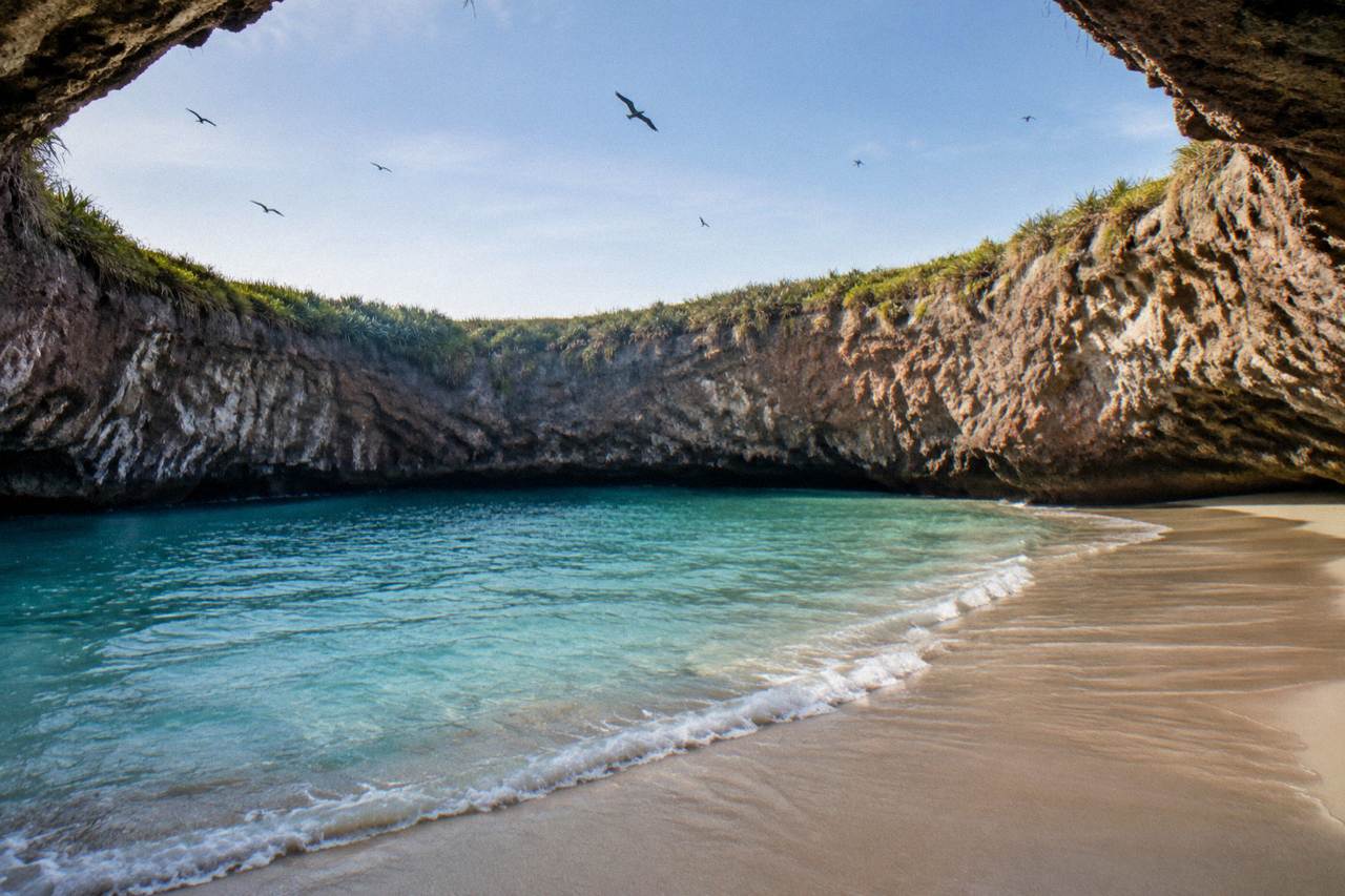A sandy cove inside a rocky crater opening to the sea, with turquoise water below