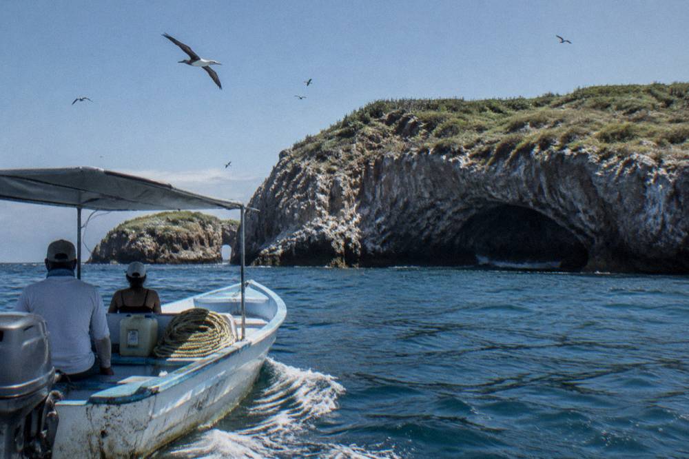 A covered small boat cruising along the cliff face of the Marietas Islands with seabirds above