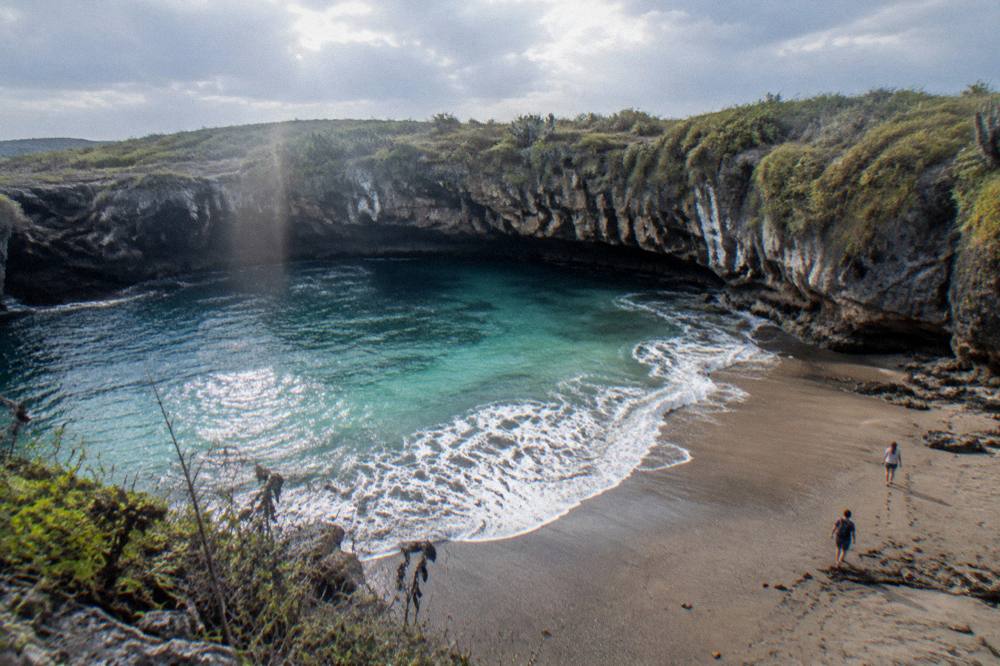 Secluded crater beach with turquoise water, vegetated cliffs, and people walking on the sand