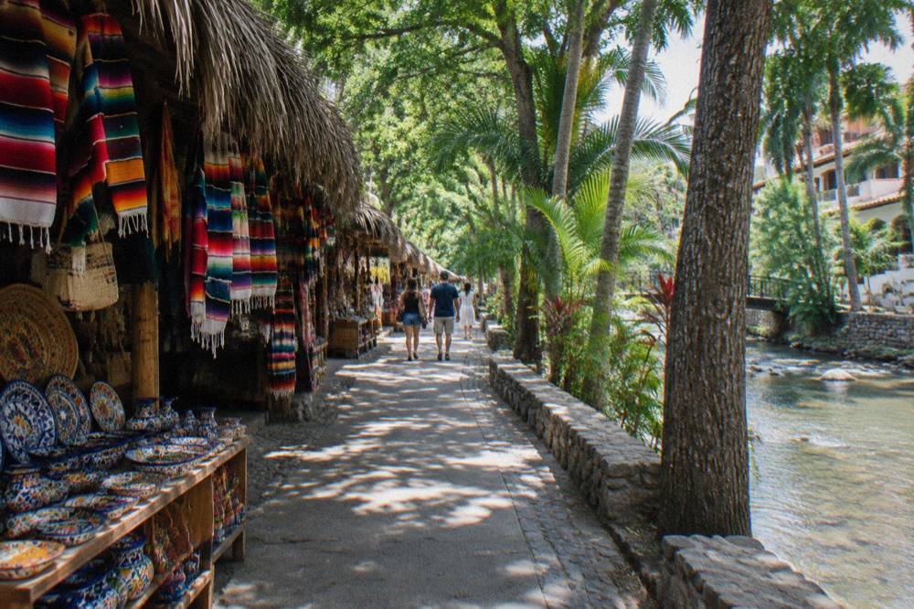Shaded riverside walkway with thatched market stalls selling blankets and pottery as people stroll