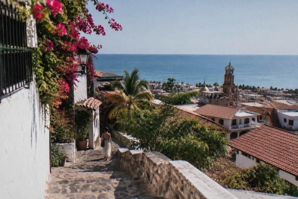 Woman walking down a cobblestone street toward a coastal town with a crowned church tower