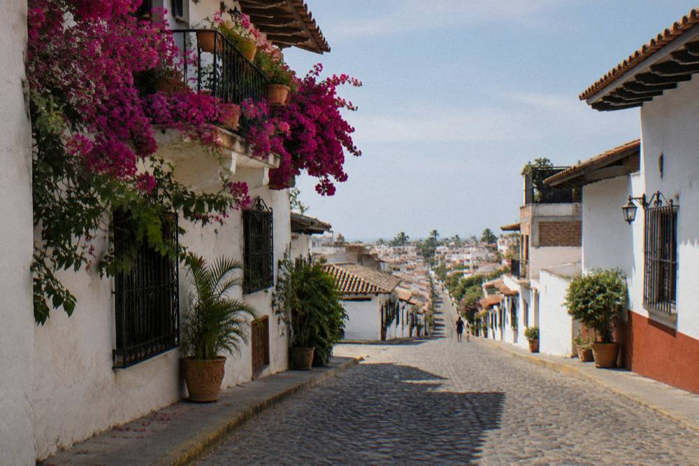 Cobblestone street lined with white colonial buildings and pink bougainvillea descending toward a distant town