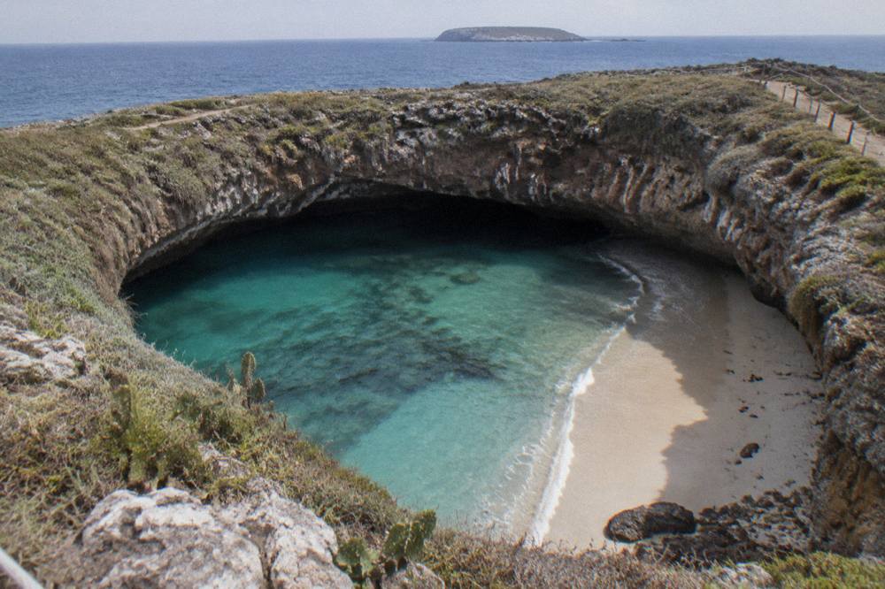 A view down into a circular crater beach with pale sand and still turquoise water below cliff walls