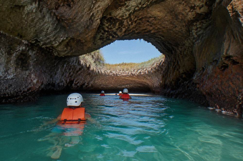 Swimmers in helmets and vests wade through a rocky sea cave toward an opening showing sky