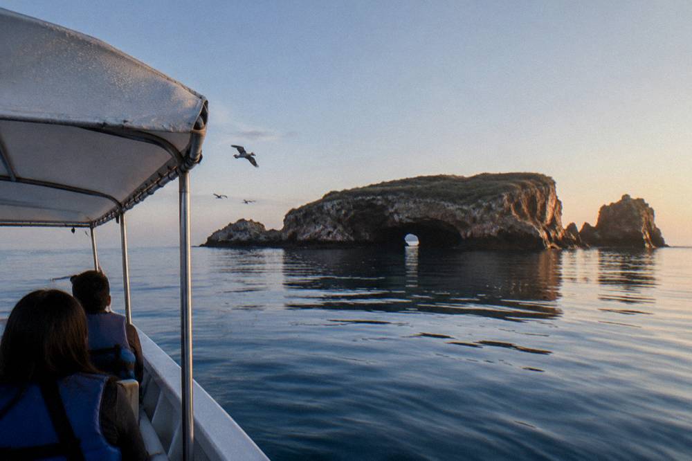 People in blue life jackets riding a shaded boat toward a rocky arch island