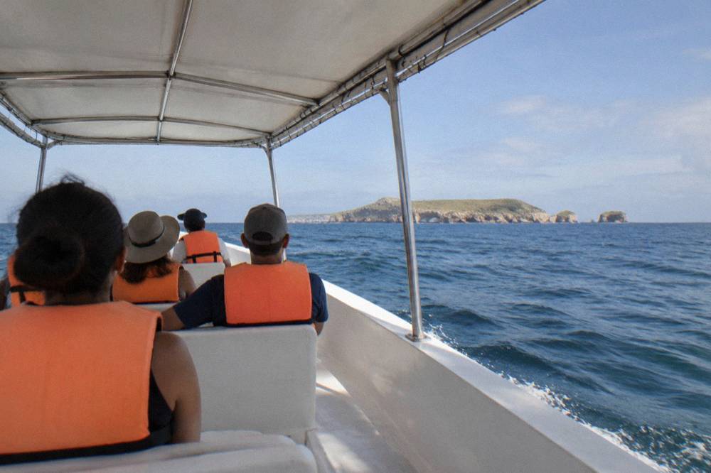 Passengers in orange life jackets riding a covered boat across blue water toward a distant island