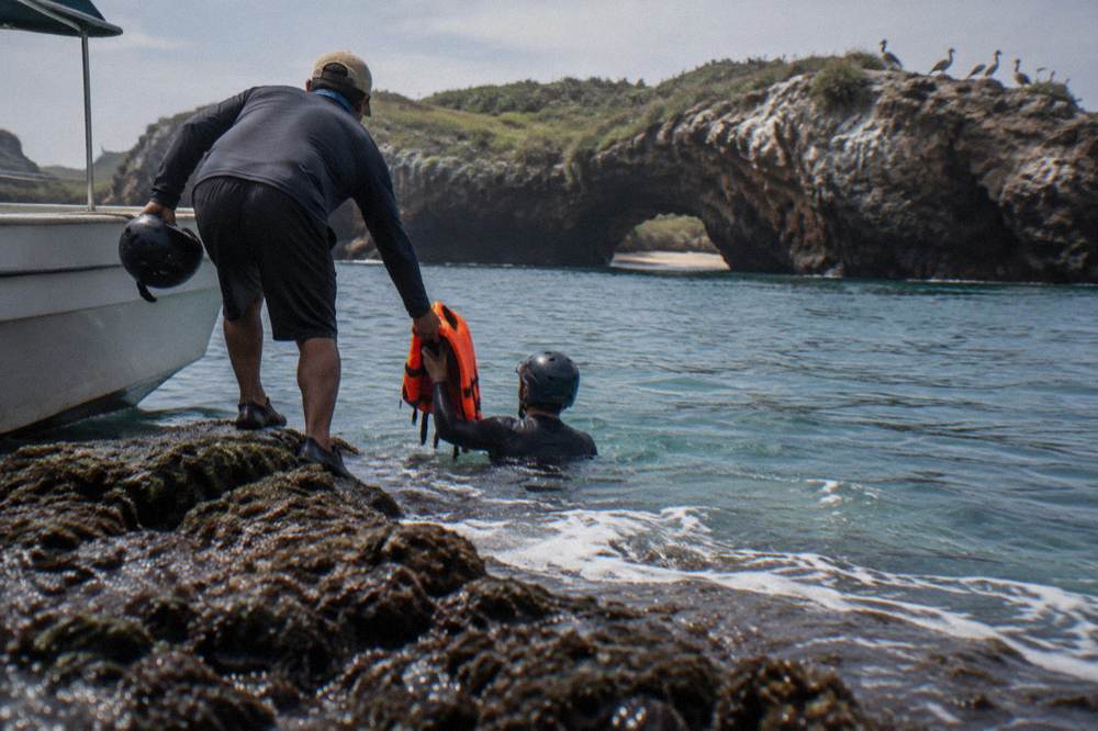 Man on rocks passing an orange life jacket to a helmeted swimmer beside a boat
