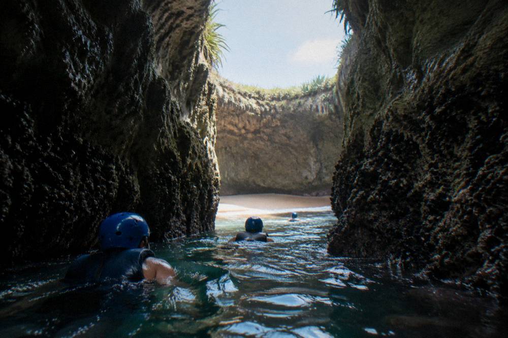 Swimmers in helmets and life vests wading through a narrow rock cave toward a hidden beach