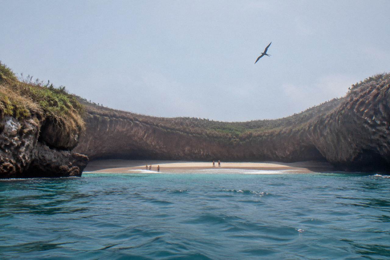 Sandy beach inside a rocky crater with people on shore and a bird flying overhead