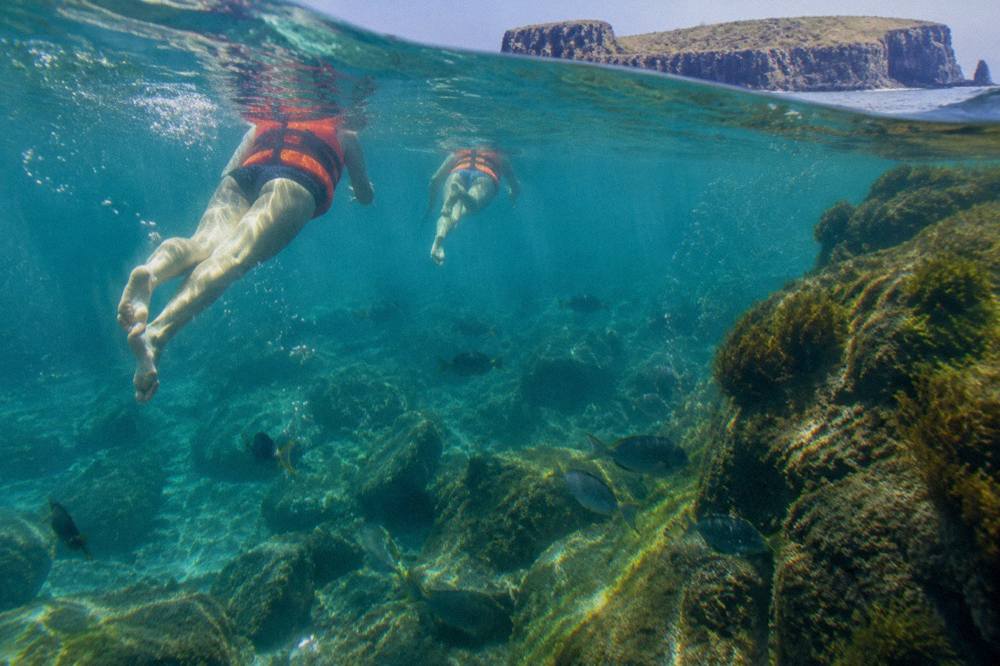 Snorkelers in the water beside a rocky shoreline with clear turquoise sea