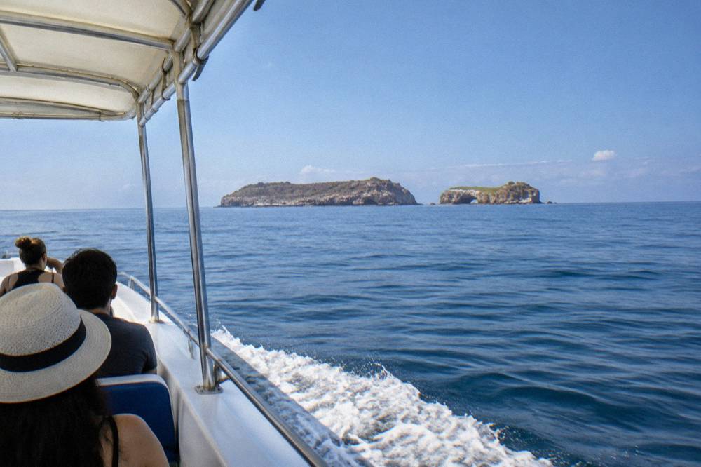 Passengers riding a covered boat across blue ocean toward distant rocky islands with an arch