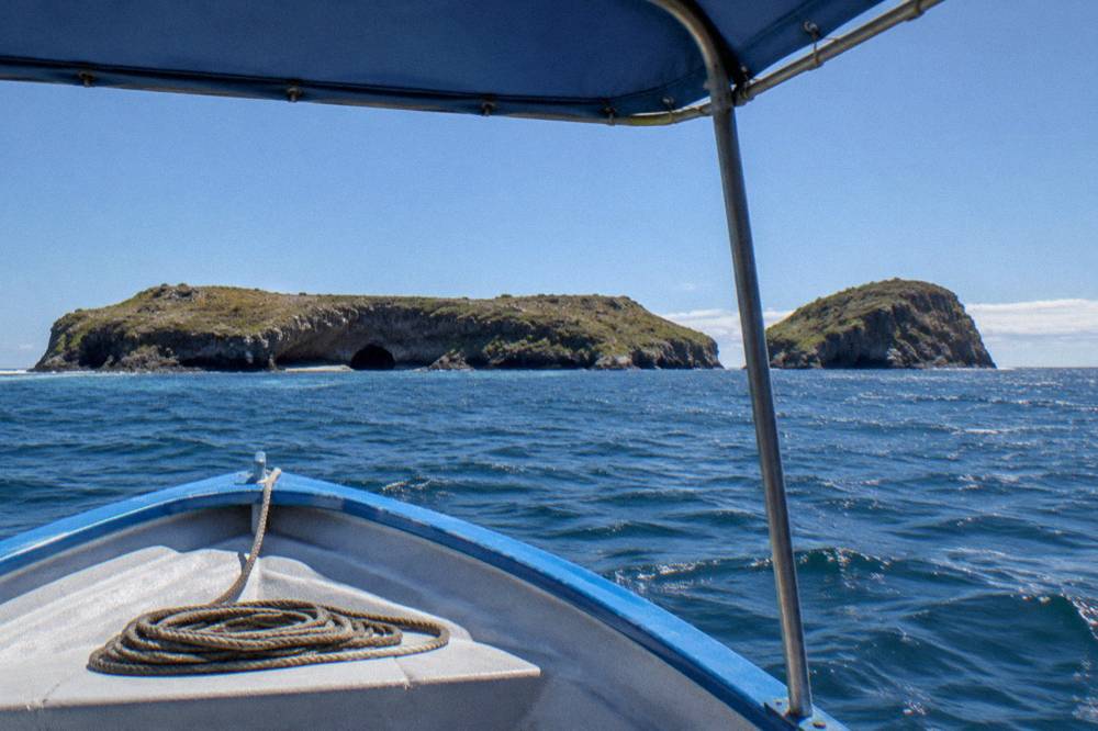 View from a boat bow over blue ocean toward distant rocky islands with a natural arch