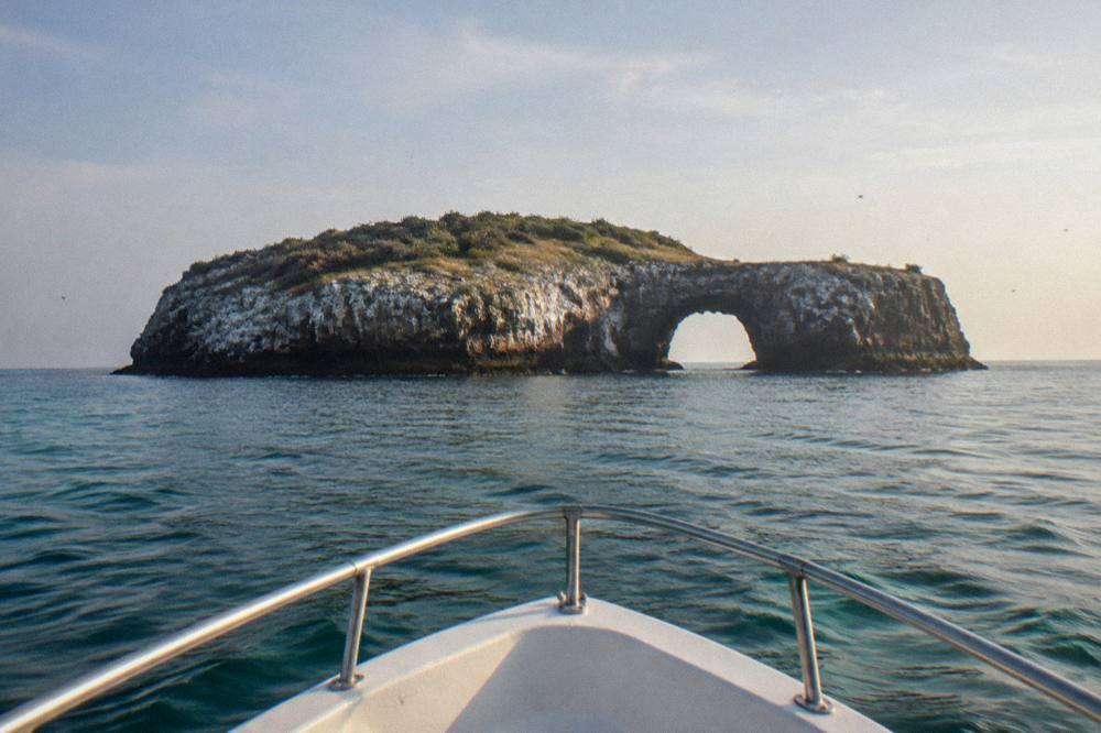 A white boat bow approaching a large rocky island with a natural arch