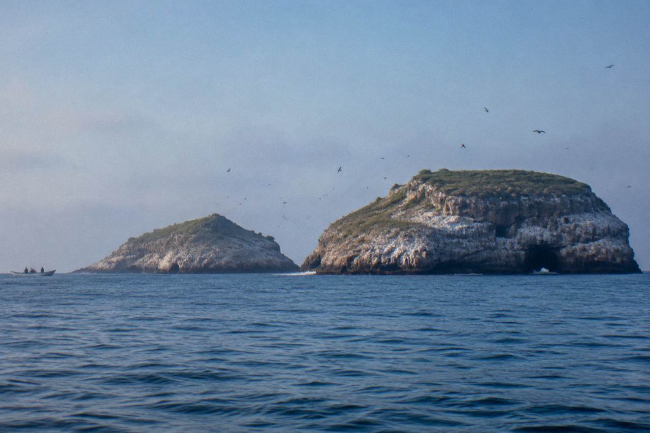 Two bird-covered rocky ocean islands under a hazy sky with a small boat nearby