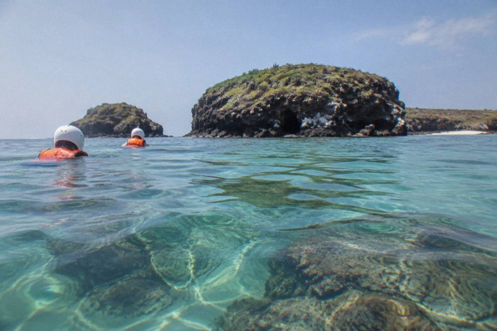 Two swimmers in helmets and life jackets float in turquoise water near green rocky islands