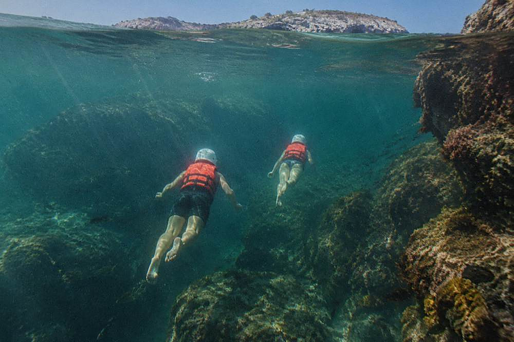 Two people in red life jackets swimming in clear green water near underwater rocks