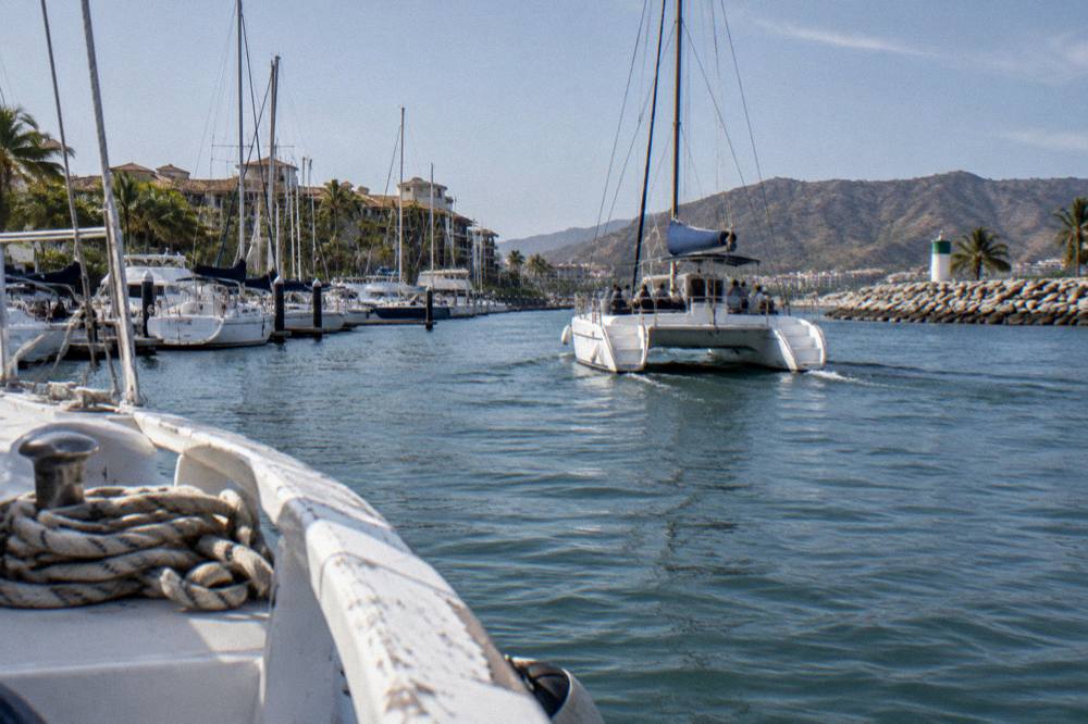 Catamaran sailing through a marina channel past docked sailboats, palm trees and resort buildings