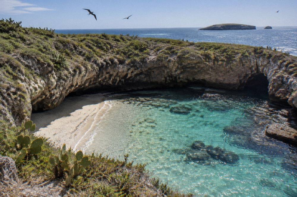 Sandy beach inside a rocky crater with clear turquoise water and seabirds overhead