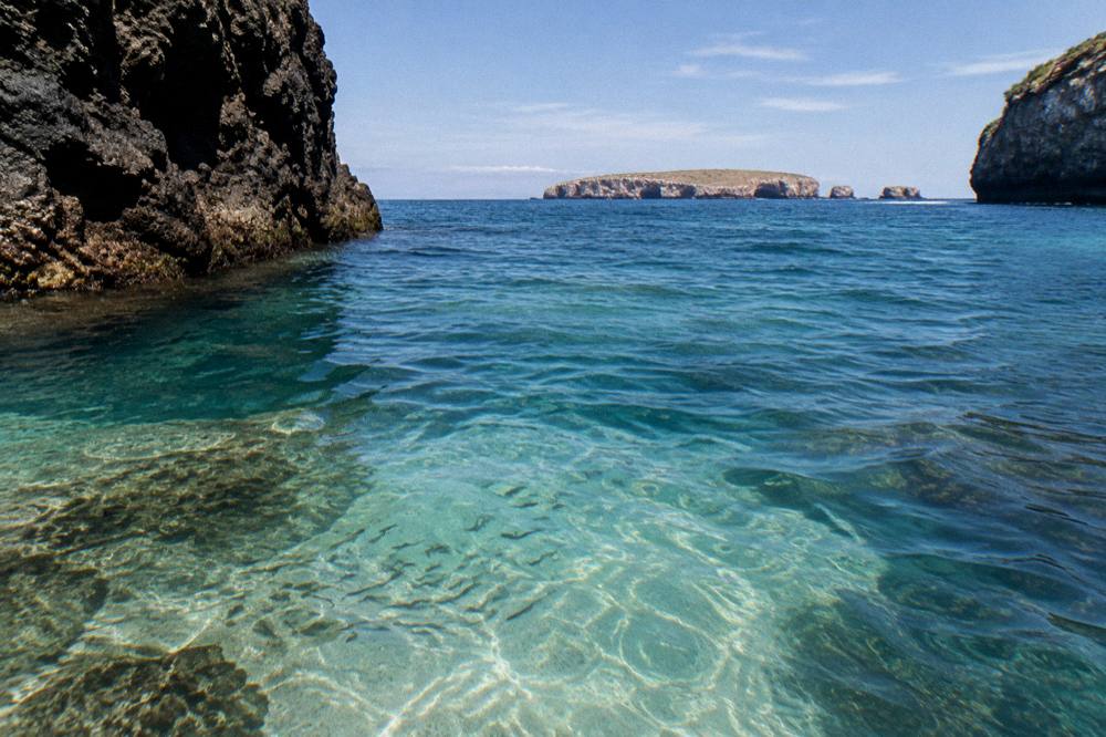 Clear turquoise water between dark rocky cliffs with distant islands on the horizon