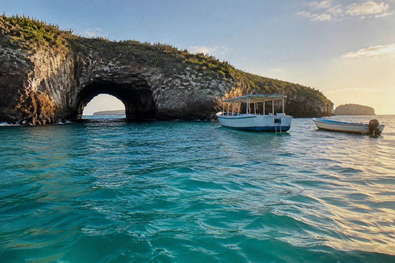 Two boats on turquoise water beside a cactus-covered rock arch island