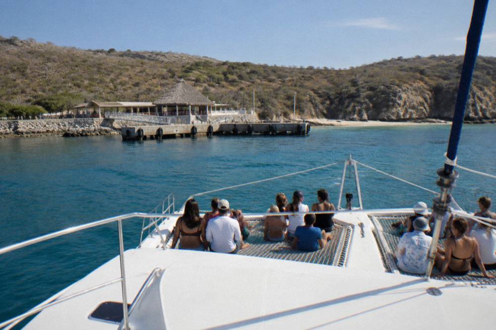 Tourists seated on a boat bow approaching a concrete pier with a thatched-roof building