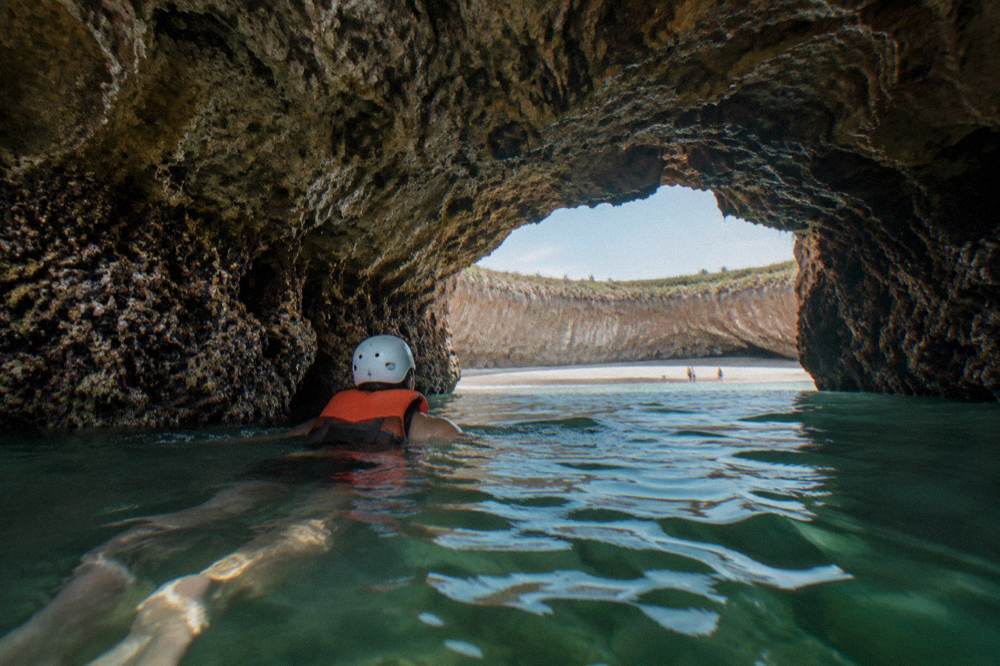 A swimmer in a helmet and life vest passing through a rock passage toward a beach