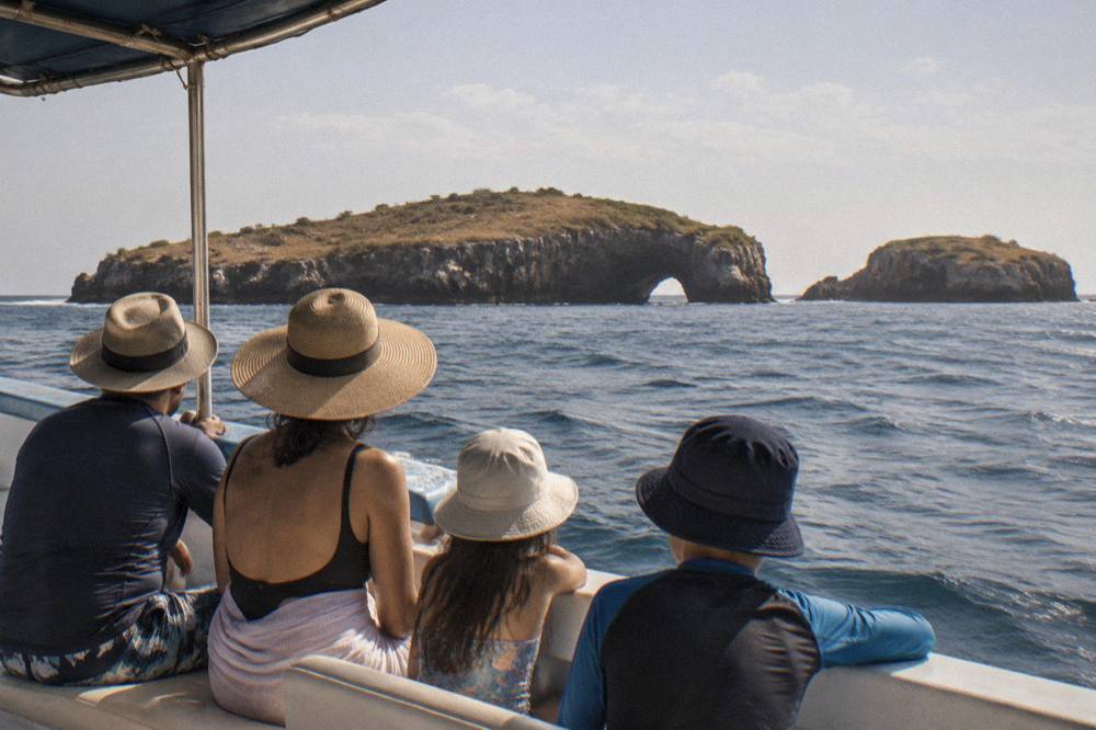 People in sun hats on a boat looking toward a rocky island with a sea arch