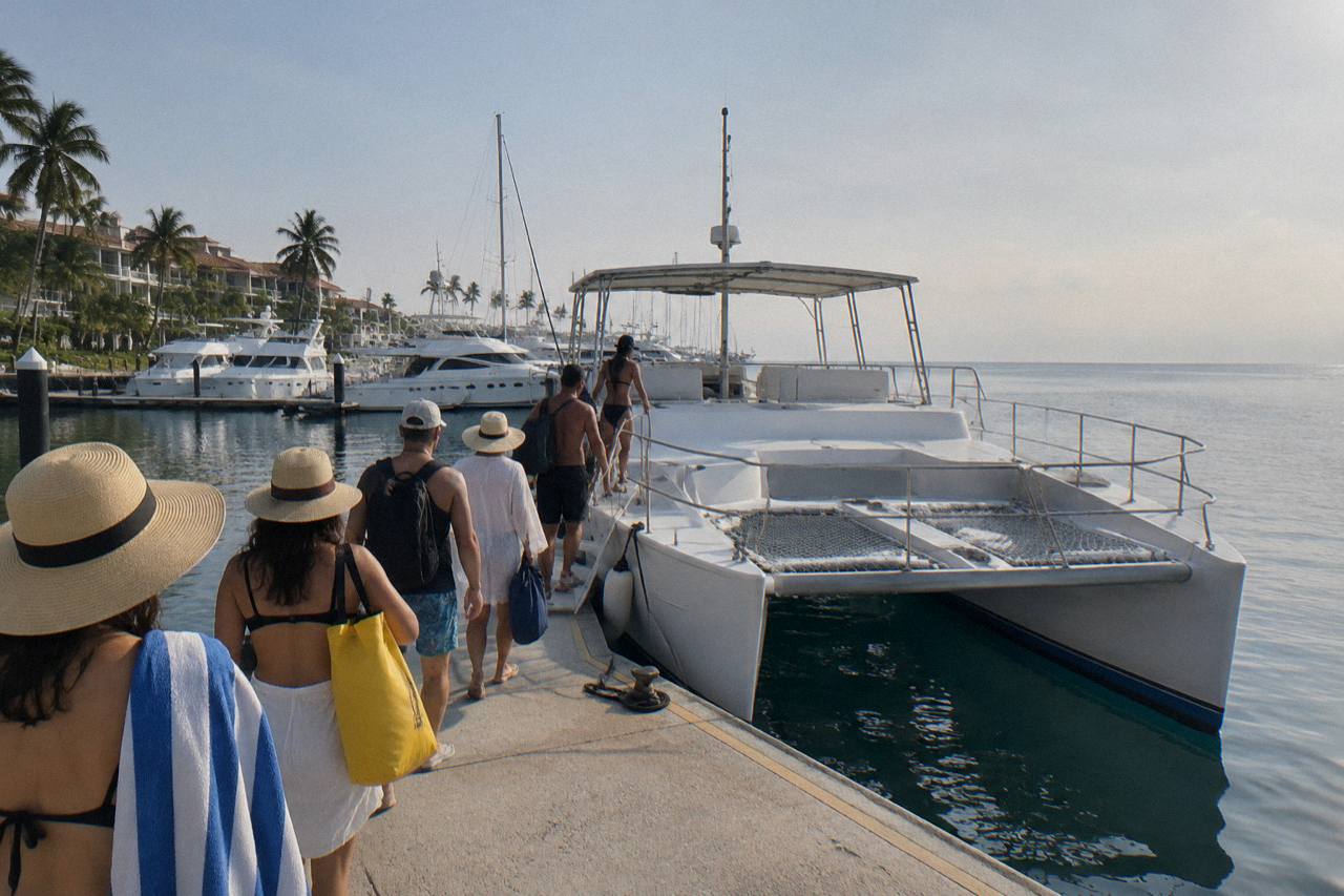 People in sun hats boarding a white catamaran at a marina with yachts and palm trees