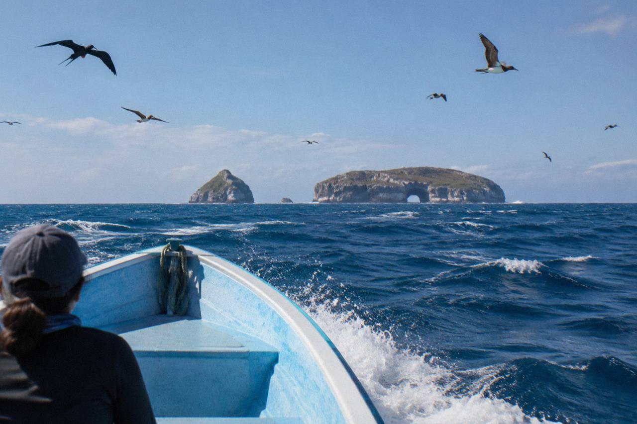 Boat bow speeding across choppy blue sea toward rocky islands with a natural arch